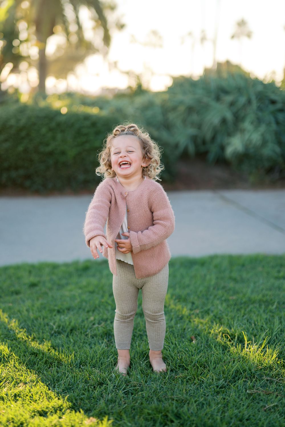 A toddler in pink sweater and khaki pants plays on green grass during golden hour.