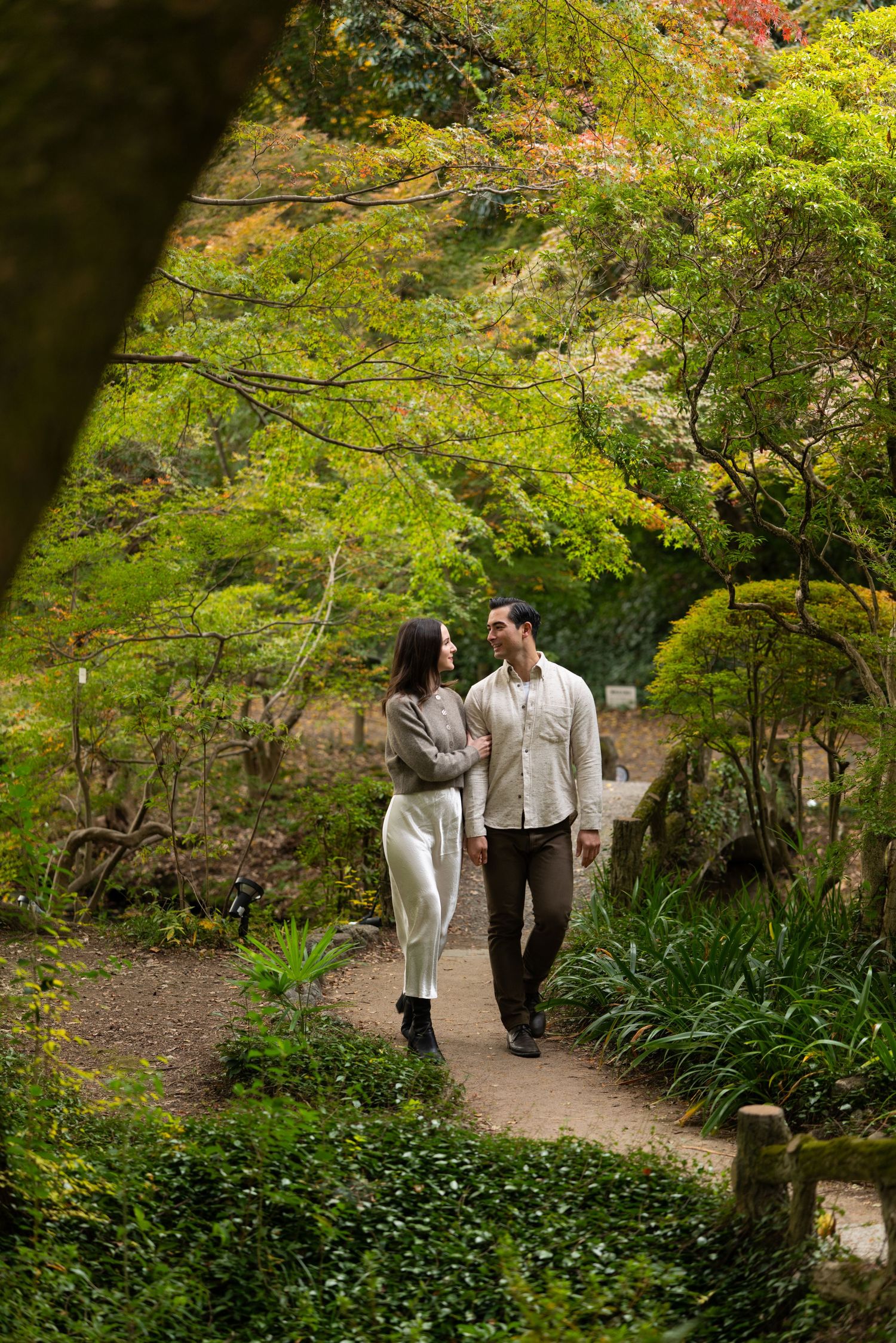 Couple walking hand in hand along a lush garden path surrounded by vibrant green and autumn foliage.