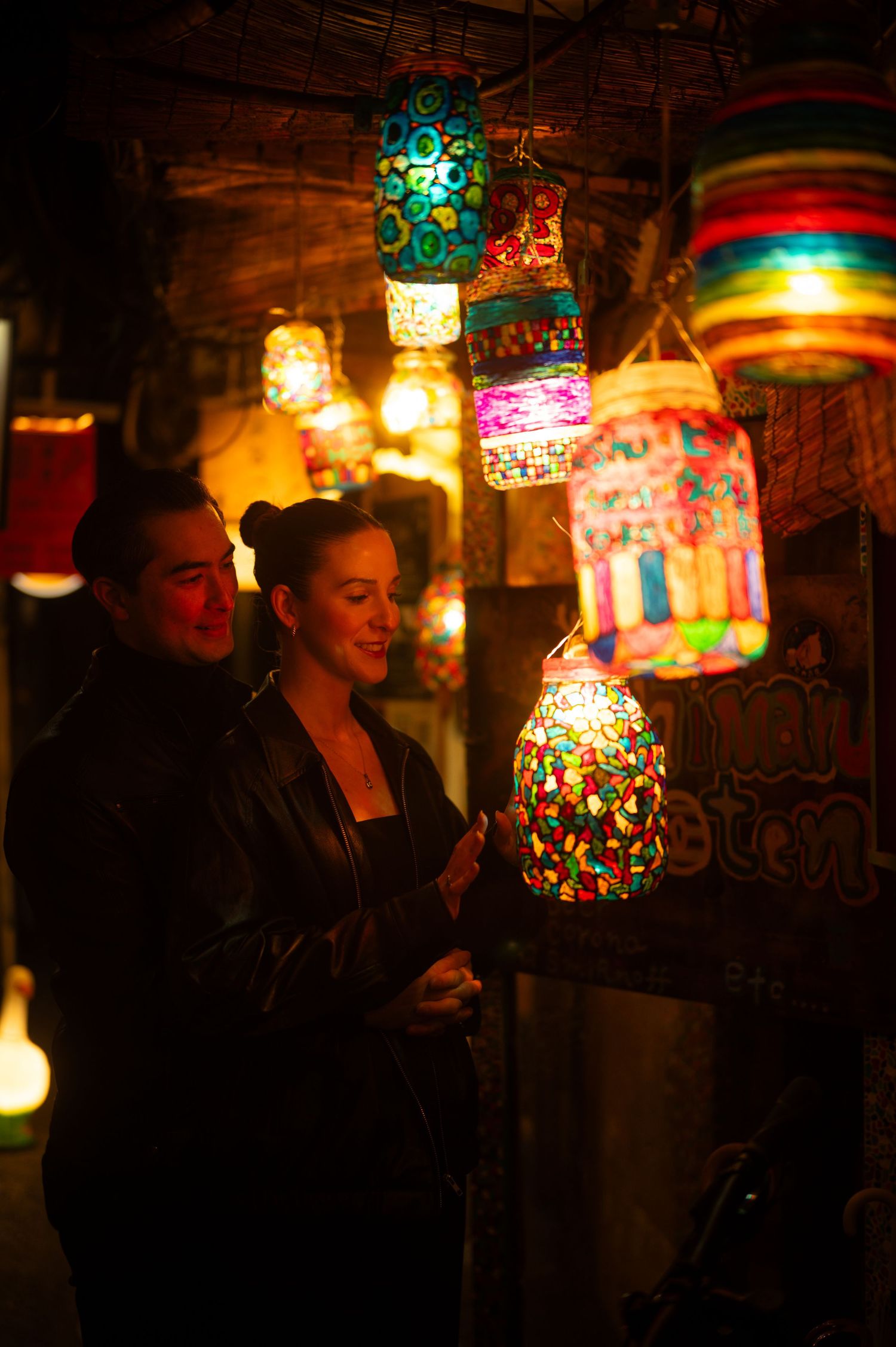 Couple admiring colorful mosaic hanging lanterns in a warmly lit market stall at night.