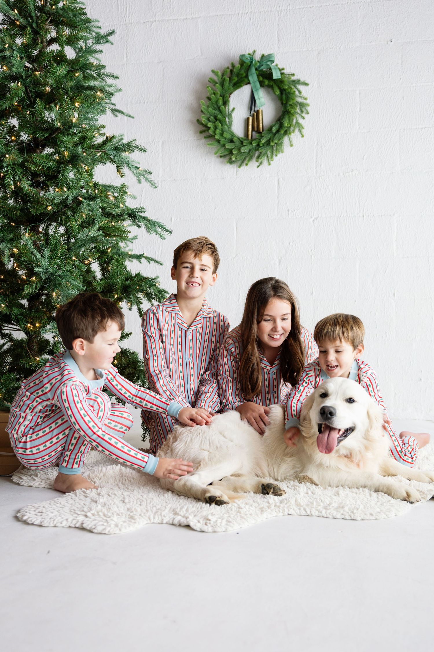 A family wearing matching red and white striped pajamas poses on a white rug with their fluffy white dog for holiday photos.