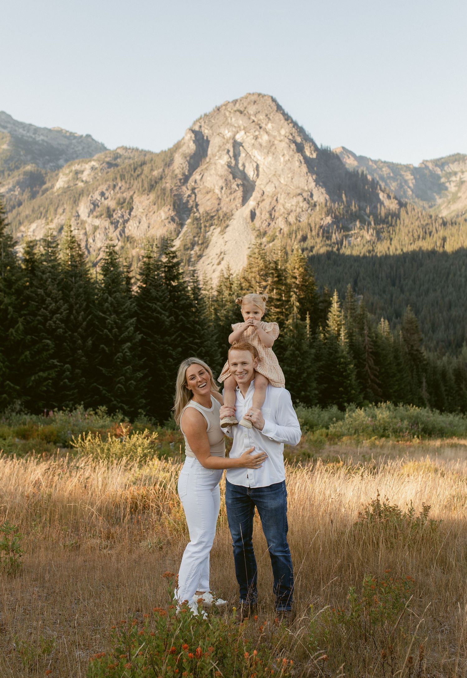 A couple shares an intimate moment in a meadow with majestic mountain peaks and evergreen forest in the background.