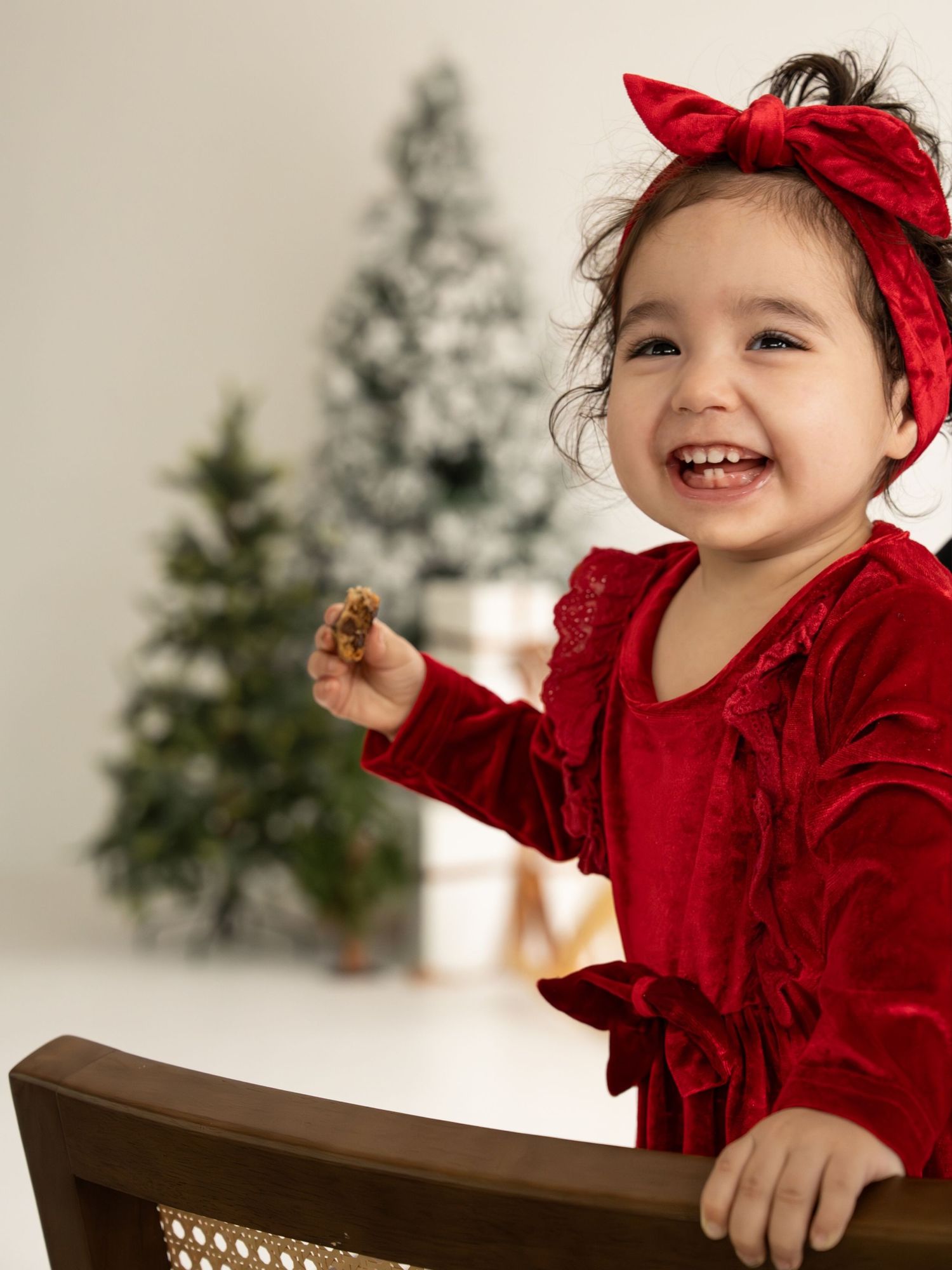 A child in a red velvet dress and bow headband smiles joyfully while playing near a Christmas tree.