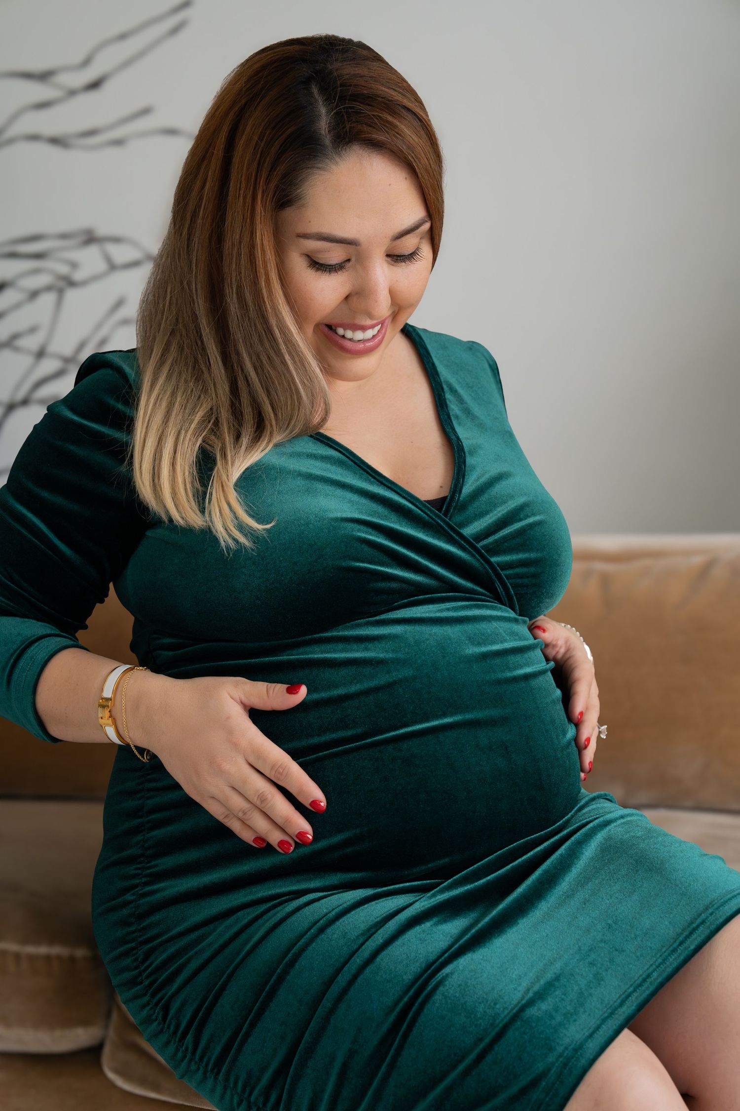 Person wearing emerald velvet dress touches their belly while sitting on couch during maternity photoshoot.