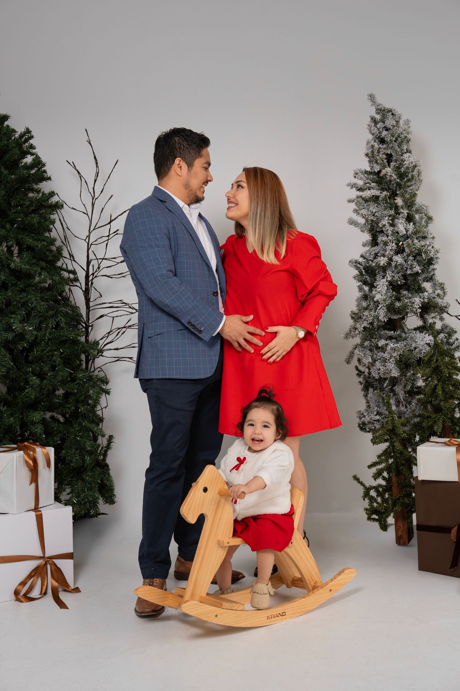 Family enjoying a holiday photoshoot with a wooden rocking horse and evergreen trees in a studio setting.