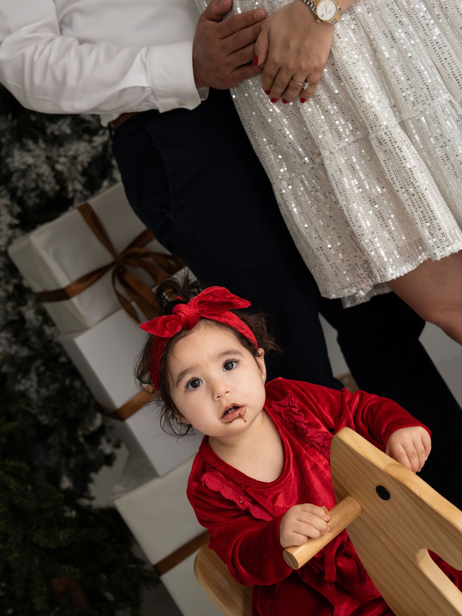 A baby in a red velvet dress and bow sits in a wooden chair against a dark background with sparkly lights.