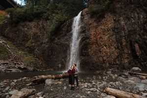 Two figures stand beneath a towering waterfall cascading down reddish-brown cliffs in a forested area.