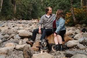 A couple sits together on large rocks by a flowing river surrounded by autumn foliage in a forest setting.