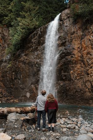 A couple admires a majestic waterfall cascading down rocky cliffs into a turquoise pool surrounded by stones.