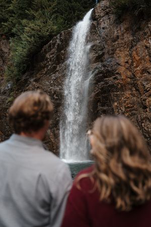 Two people stand together viewing a tall waterfall flowing down orange-hued cliffs in a forest setting.