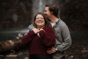 Two people embrace and laugh together near a scenic waterfall on an autumn day.