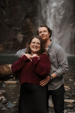 A romantic couple embraces in front of a waterfall, wearing burgundy and gray sweaters.