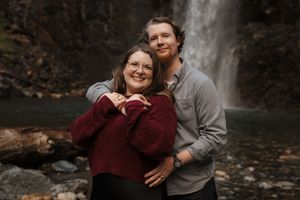 A romantic engagement photo shows a couple sharing joyful moments by a forest waterfall.