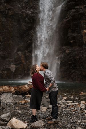 A couple shares an intimate moment by a majestic waterfall on a rocky shore surrounded by natural beauty.