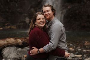 A couple embraces while sitting on fallen logs near a misty waterfall in the background.