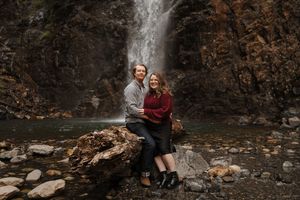 Two people share an intimate moment by a waterfall surrounded by fallen autumn leaves and rocks.