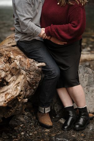 Close-up view of two people sitting together on a fallen log wearing burgundy and gray sweaters near a waterfall.