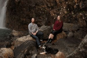 Two people sit on rocky outcropping near a waterfall in nature during an outdoor photo session.