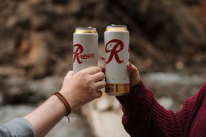Two hands holding beer cans together in a celebratory cheers gesture against a blurred outdoor background.