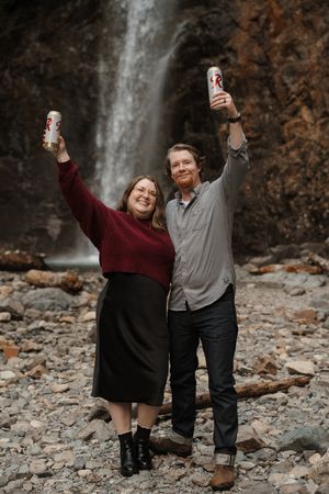 A couple celebrates with drinks near a scenic waterfall surrounded by rocky terrain and autumn foliage.