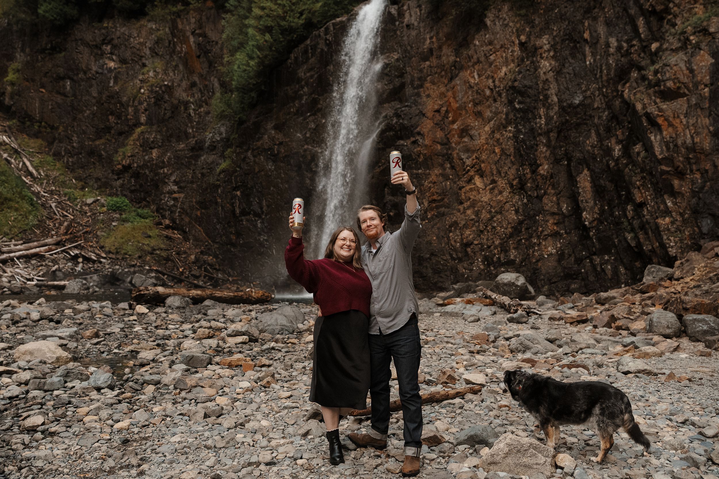 A couple walks their dog along a rocky creek bed with a cascading waterfall in the background.