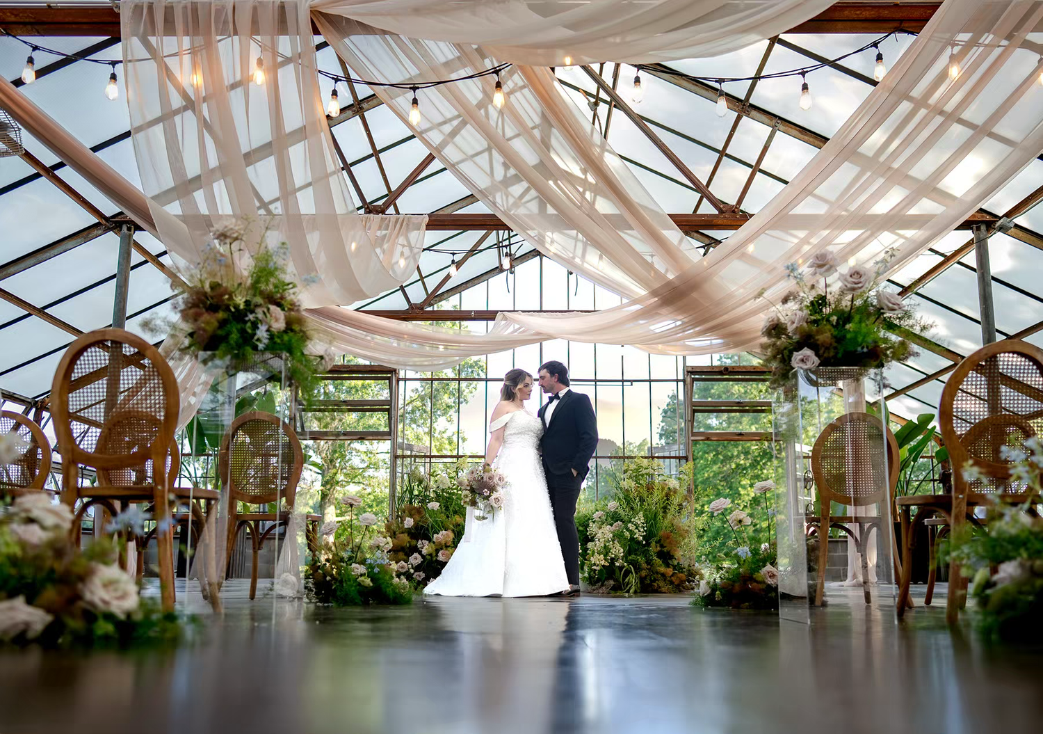 A couple celebrates their wedding ceremony in the stunning Jorgensen Farms greenhouse venue with draped fabric and floral arrangements.