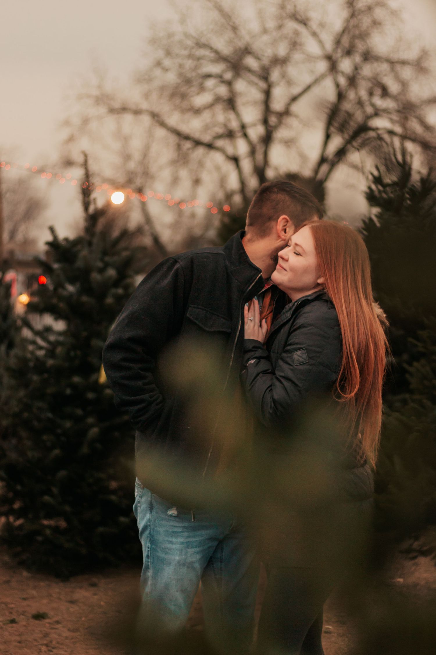 Couple sharing a romantic moment at a Christmas tree farm at dusk, surrounded by pine trees  | Westminster Colorado Proposal Photographer