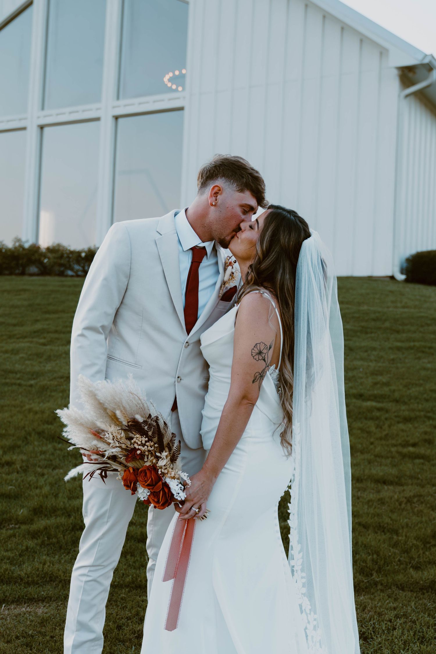 A couple shares a romantic kiss in front of a white barn on their wedding day with green grass in the background.