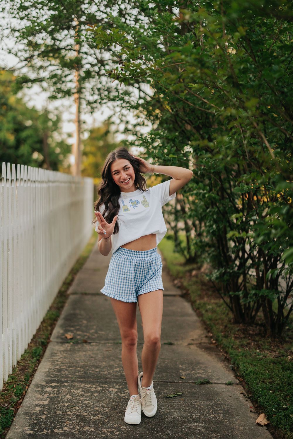 Playful outdoor poses along a white picket fence path wearing gingham shorts and a crop top.