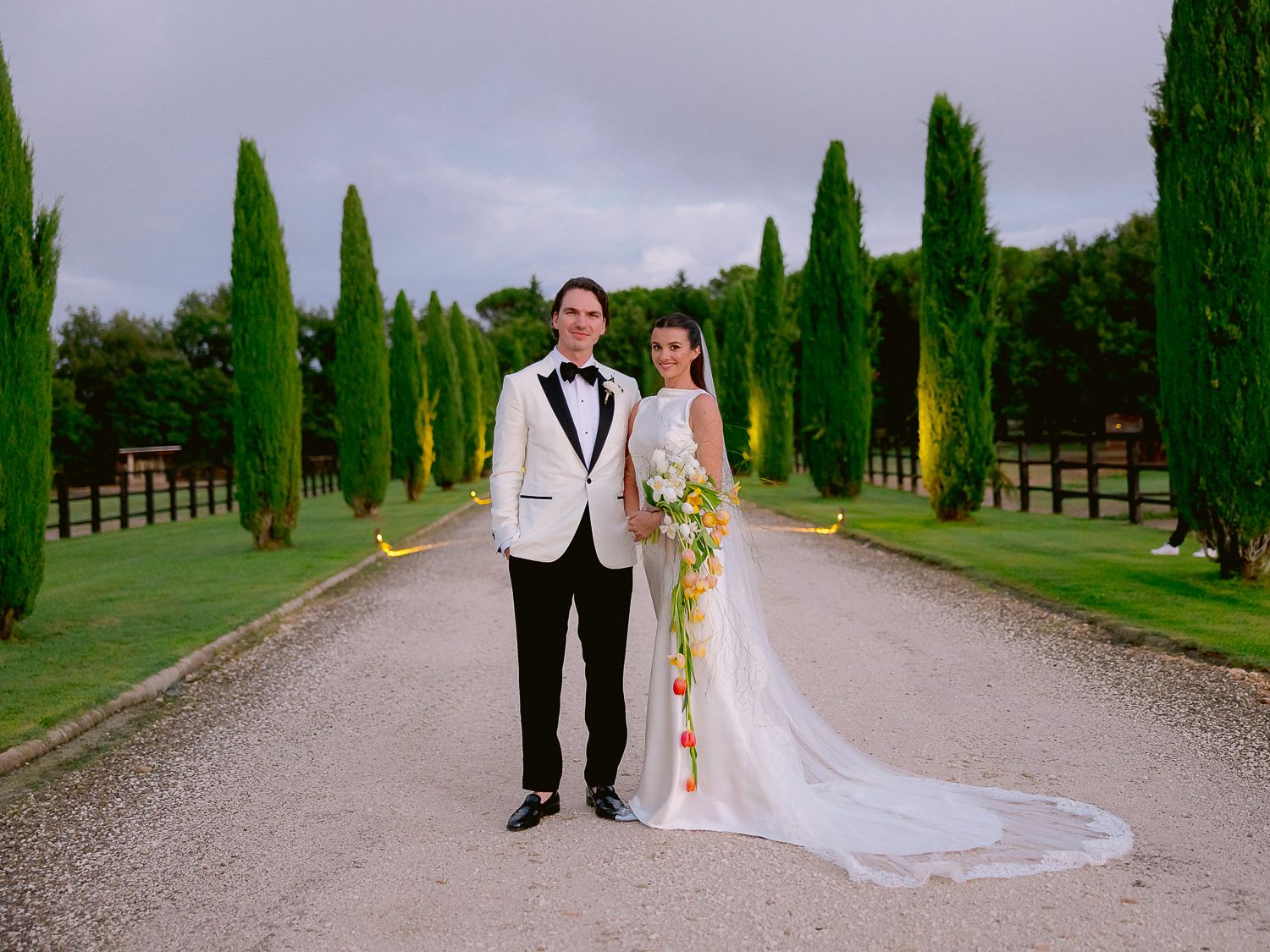 A romantic wedding couple walks down a tree-lined path with tall cypress trees creating a stunning natural aisle.
