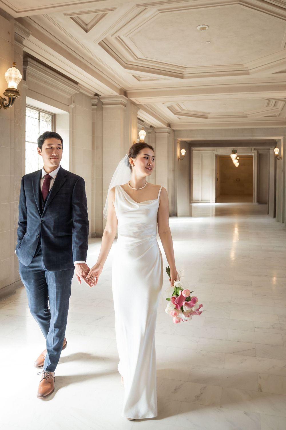 A couple in wedding attire holds hands while walking through an elegant white ballroom with ornate ceiling details.