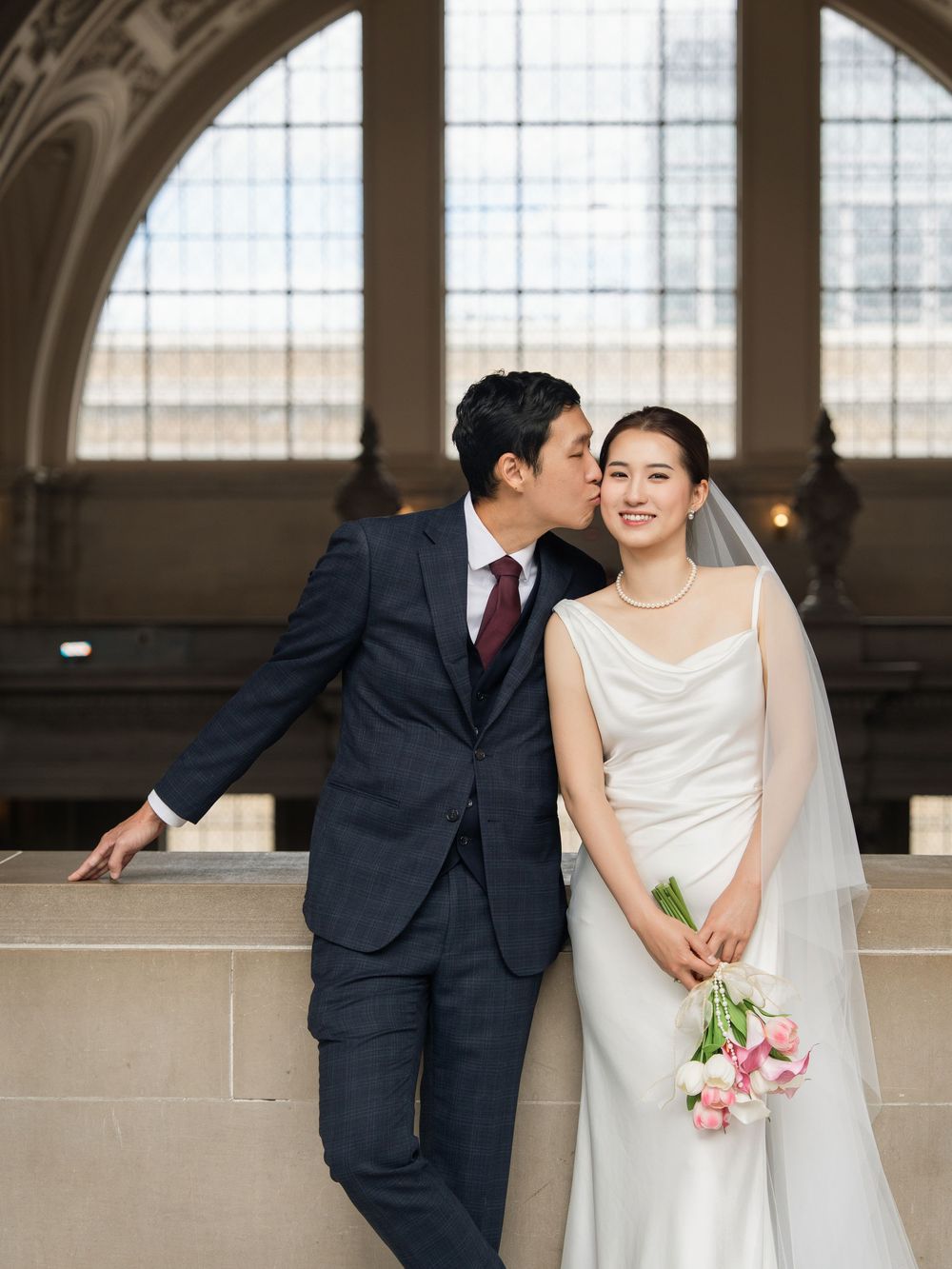 A couple shares an intimate moment during their wedding at a grand venue with arched windows and architectural details.