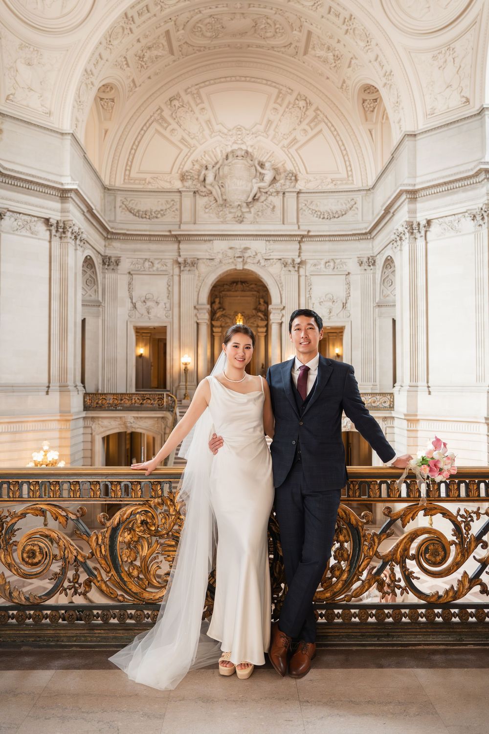 A wedding couple stands in an ornate baroque hallway with gilded railings and decorative white architecture.