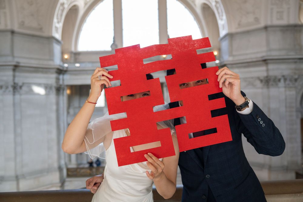 A sequence showing two people holding up red double happiness characters in front of a stone building.