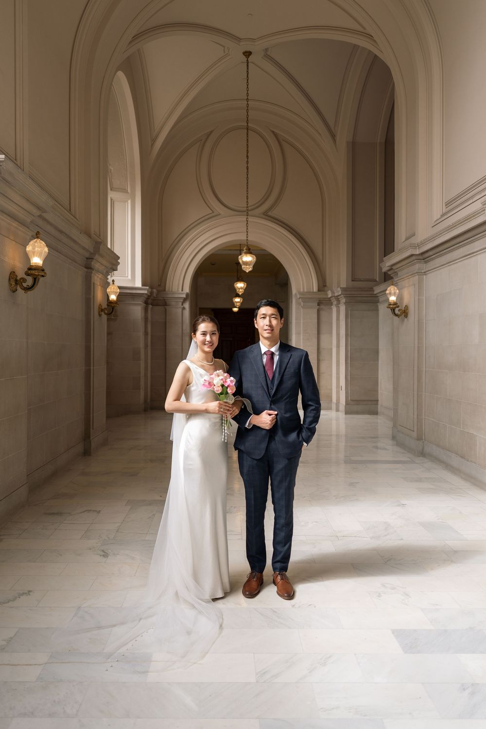A wedding couple poses playfully in a grand marble hallway with elegant architectural details and hanging lights.