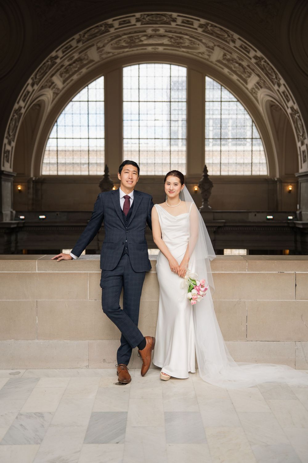 A wedding couple poses playfully in an elegant arched hallway with stone walls and natural lighting.