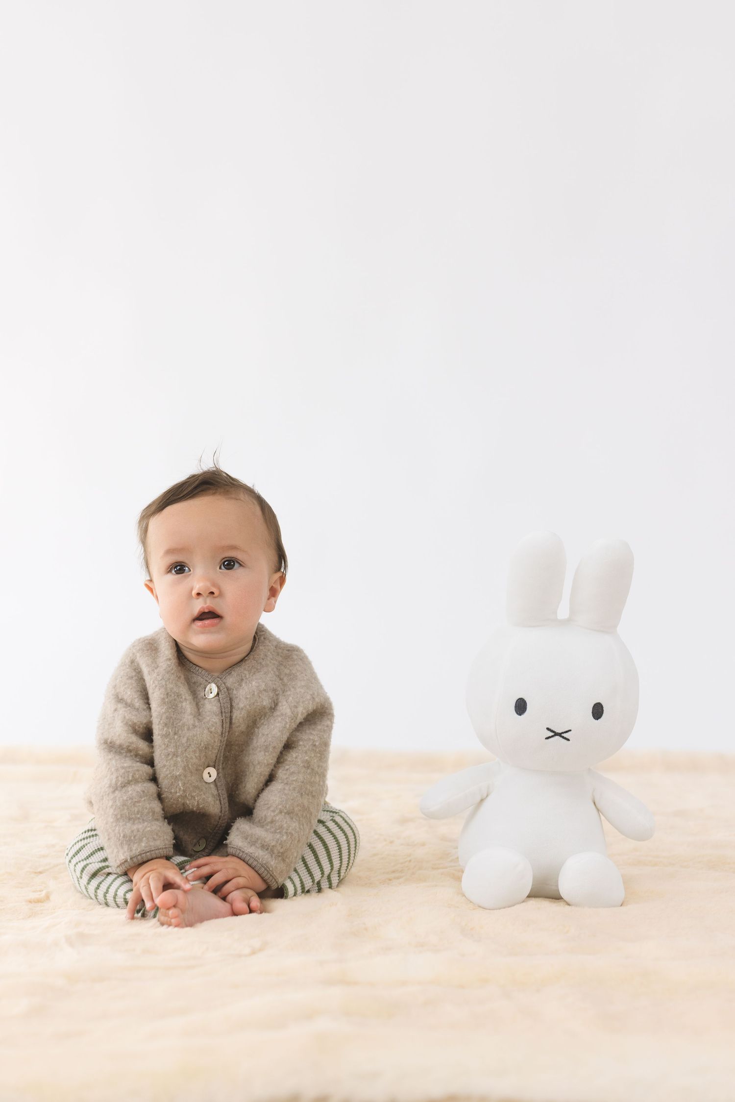 Baby in gray cardigan and striped pants sits on cream carpet next to white plush bunny toy in studio setting.