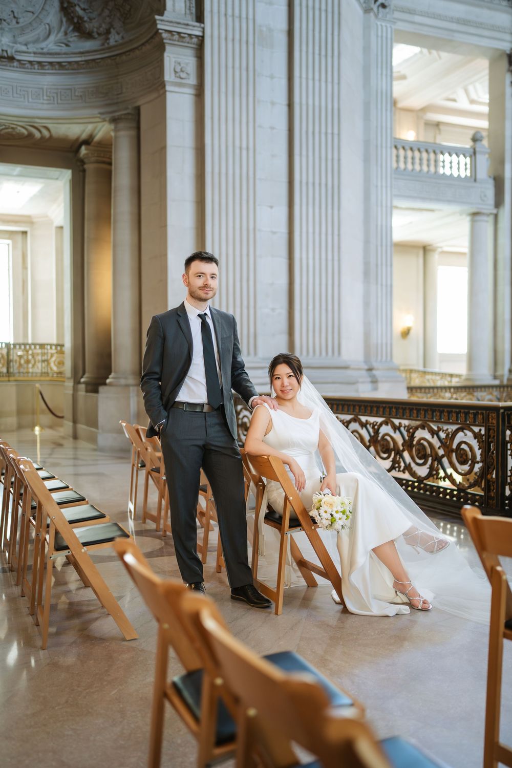 Newlyweds pose in a grand hall with wooden chairs and ornate architectural details.