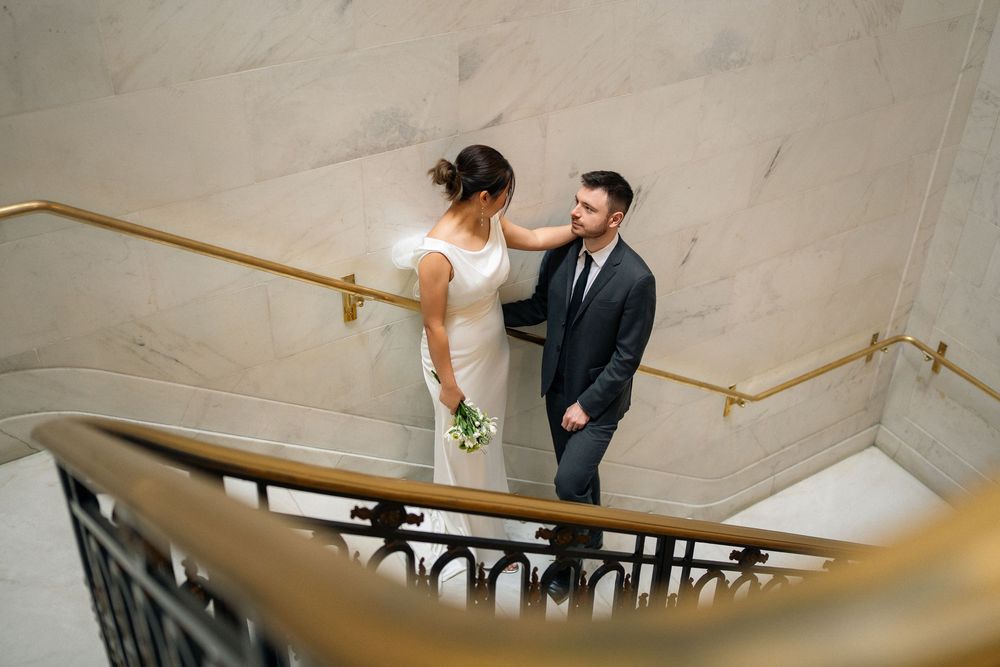 Couple poses on elegant marble staircase with gold handrail in wedding attire.