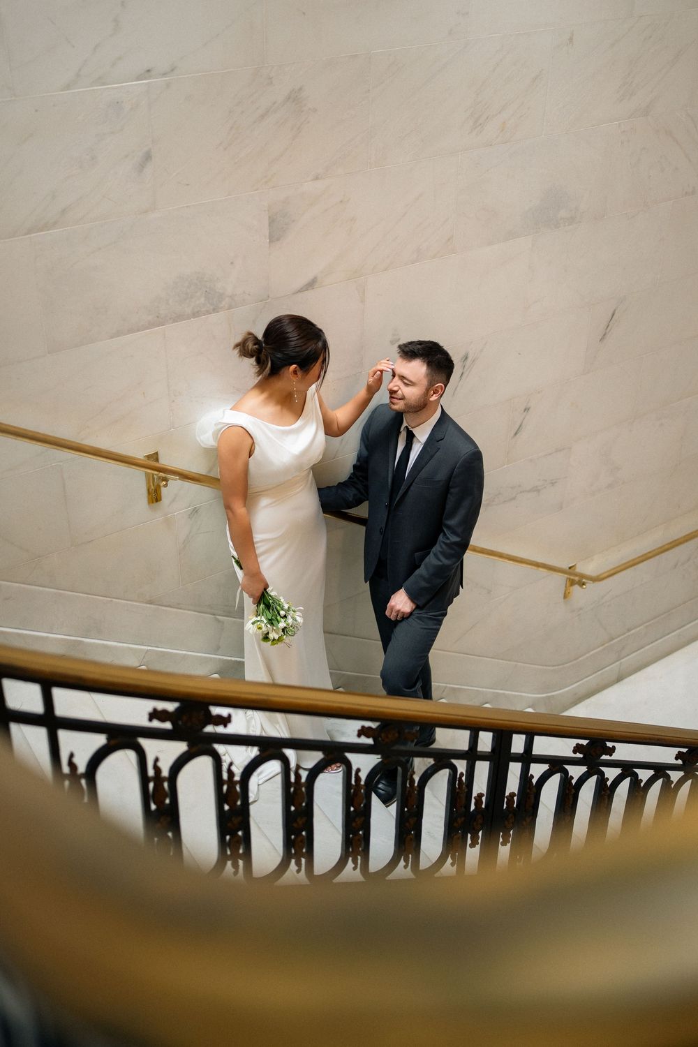 A couple poses playfully on a grand marble staircase with gold handrails.