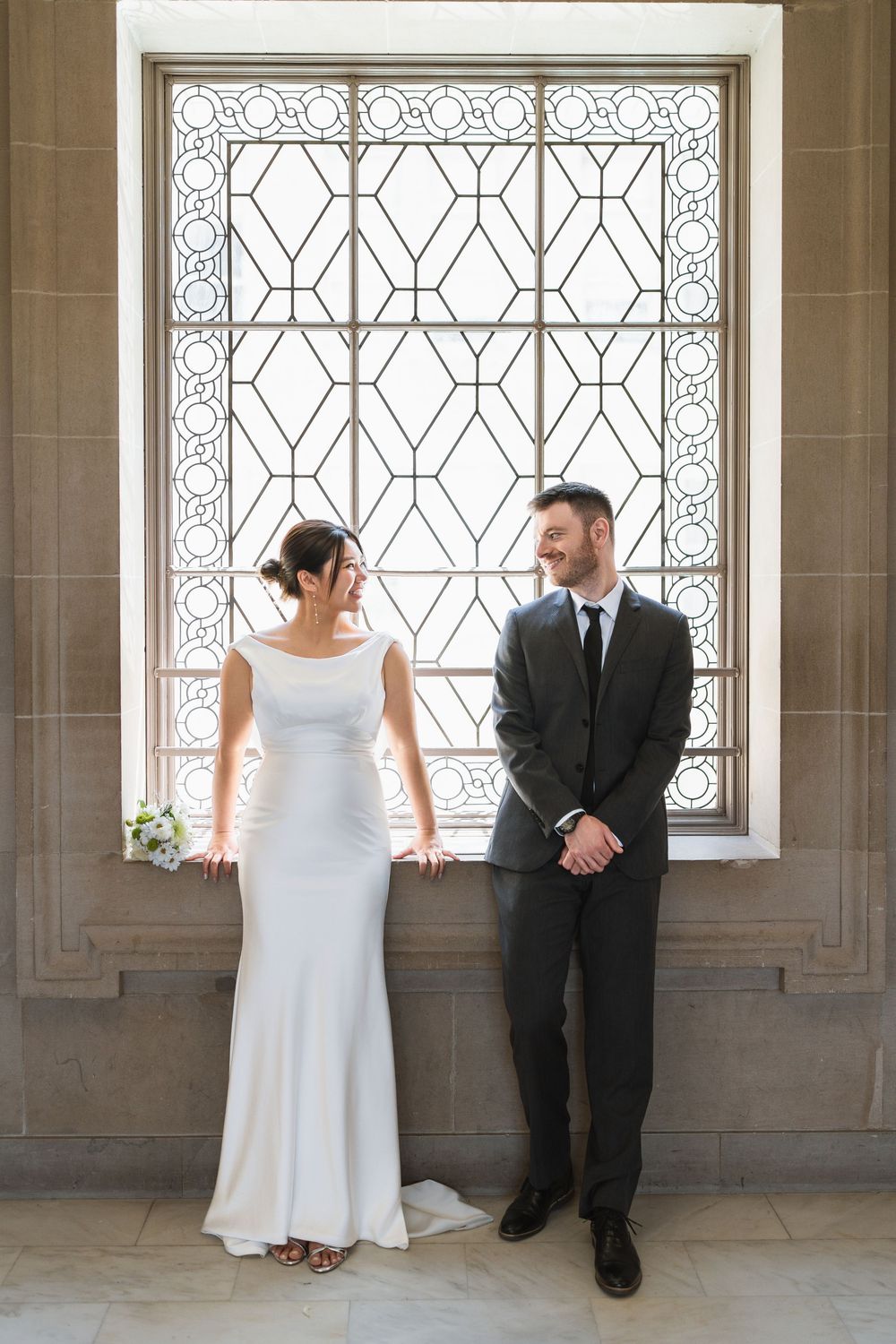 Wedding couple stands by ornate iron windows in historic building interior.