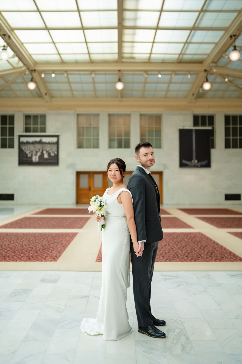 Wedding couple poses in an elegant room with skylights and patterned red carpet.