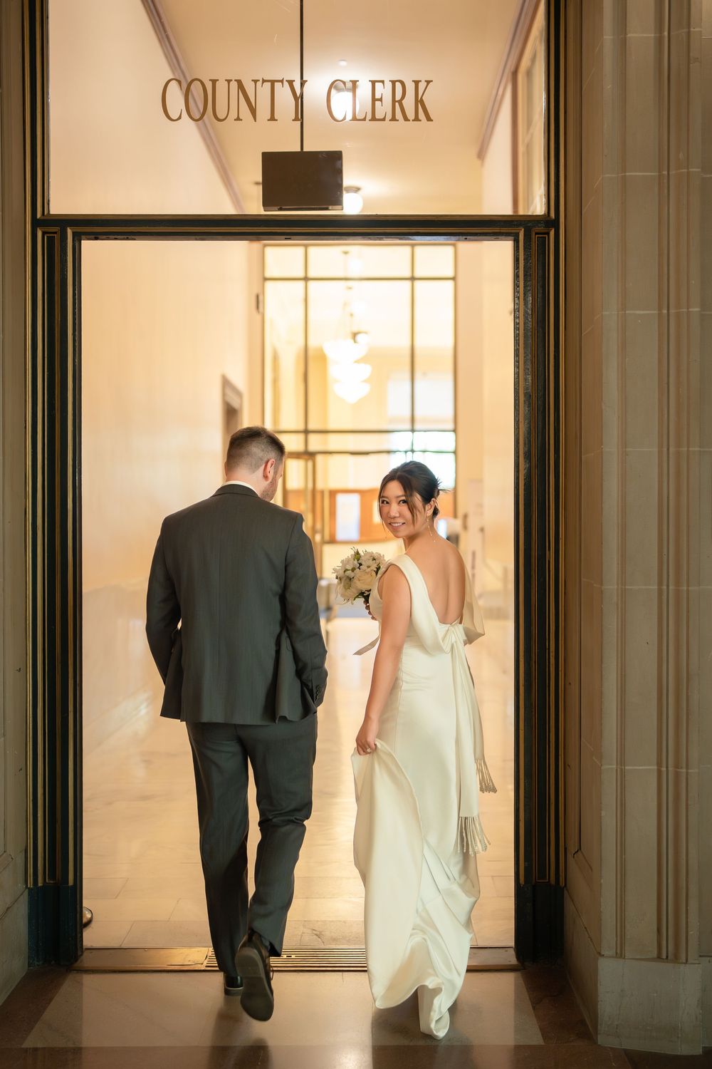 A couple walks through a brightly lit government building hallway with County Clerk signage.