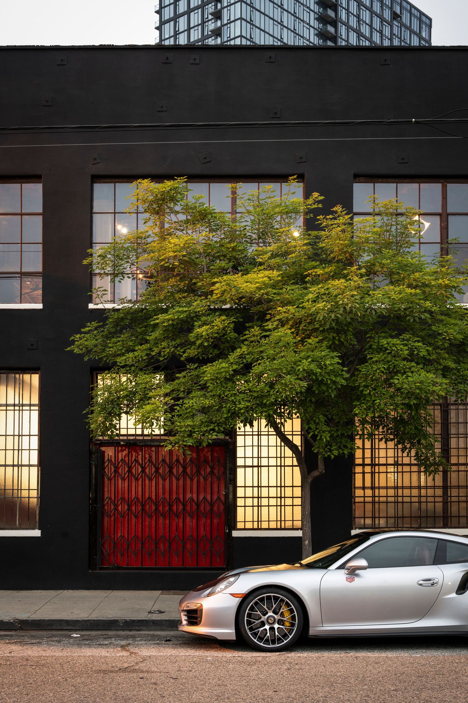 A sleek silver Porsche sports car parked in front of a modern building with red accents and green foliage.
