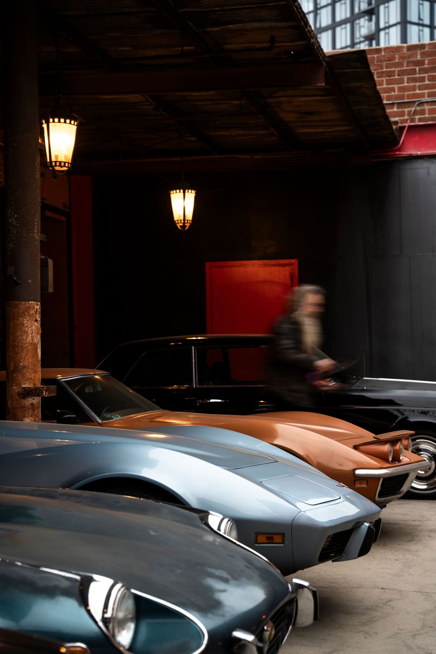 Vintage sports cars lined up in a dimly lit brick garage with atmospheric lighting and moody ambiance.