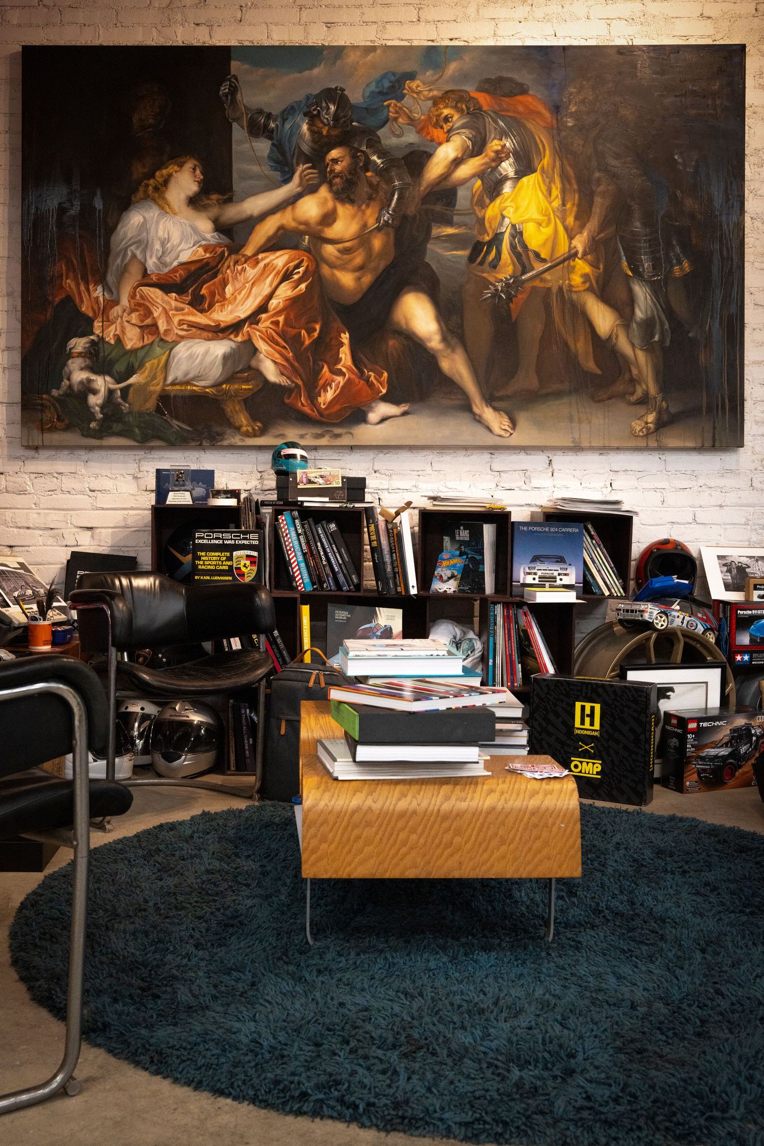 Classical art paintings adorn brick wall above cluttered office space with blue carpet and scattered furniture.