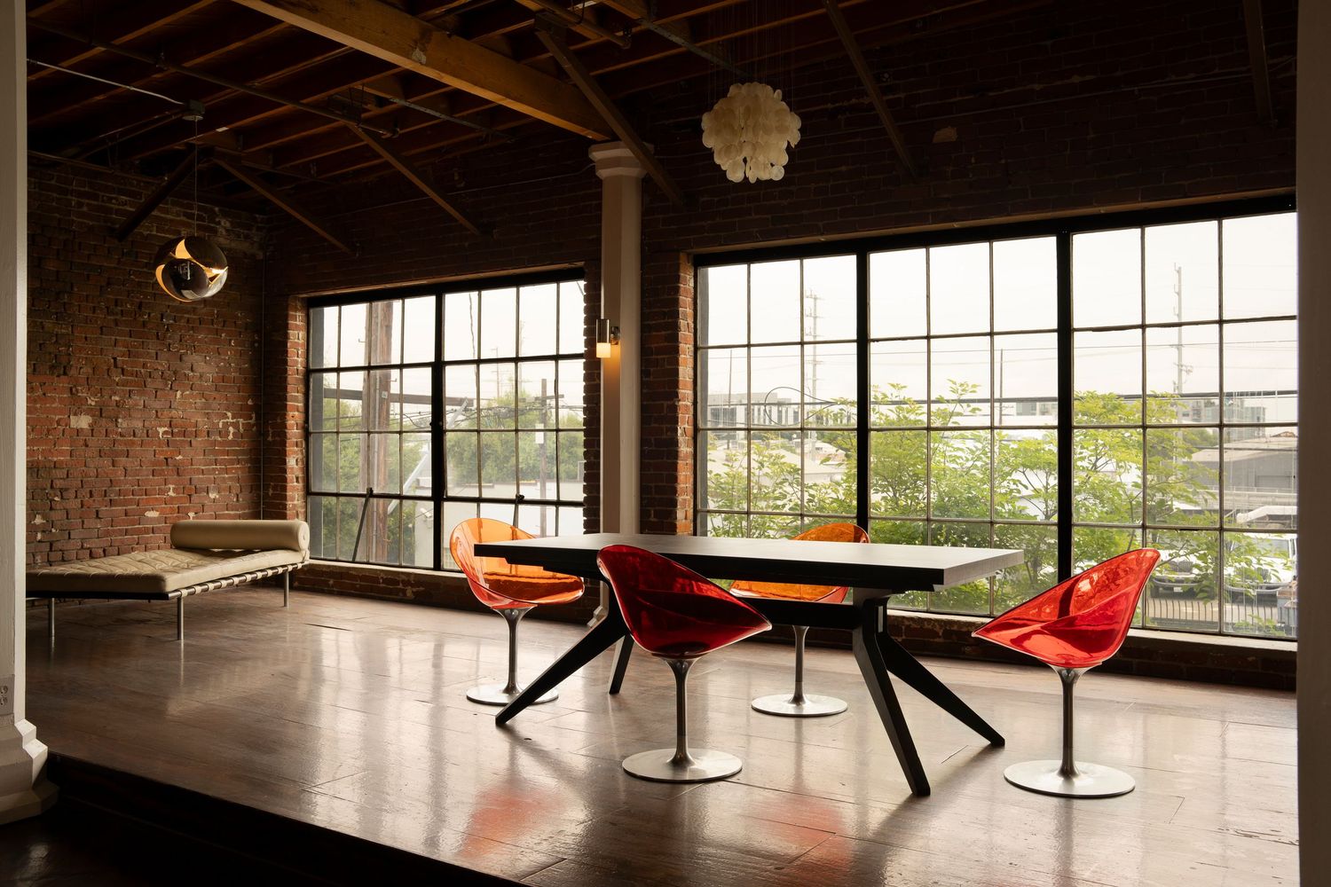Modern industrial loft space with large windows, exposed brick walls, and red retro dining chairs around black table.