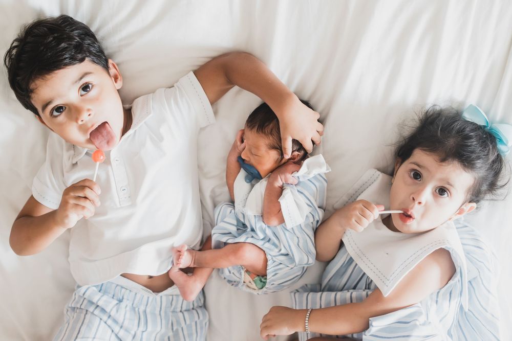 Three siblings in white clothing lying together on a bed while eating popsicles and making silly expressions.