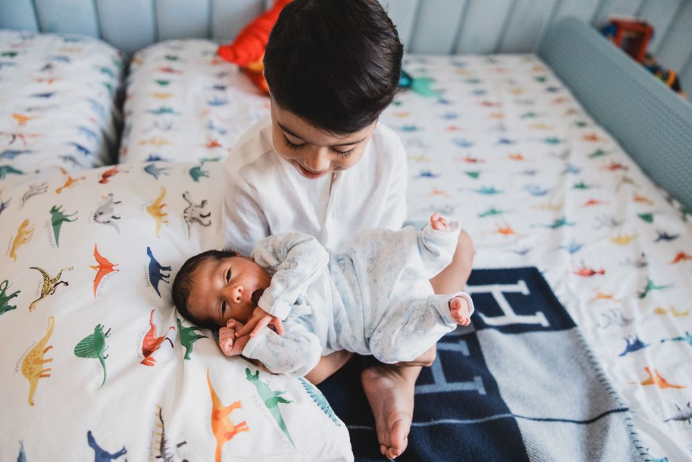 Two children in white clothing play and laugh together on a colorful dinosaur print bedspread.