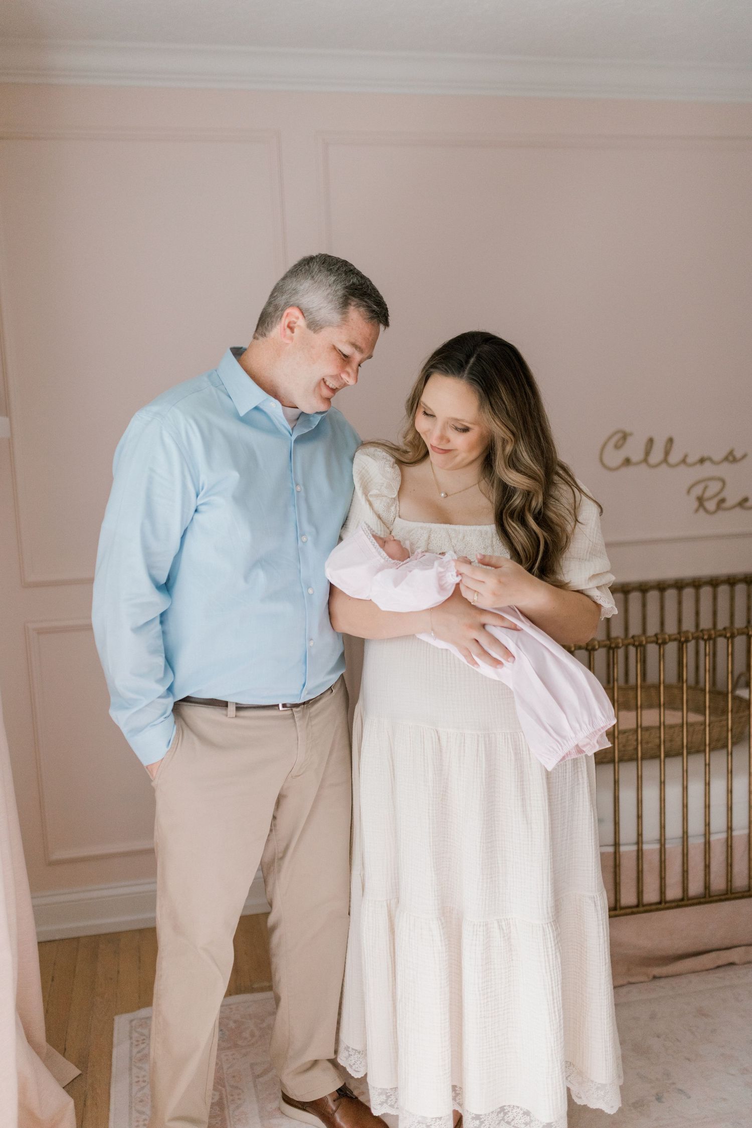 A couple in coordinating light blue and white outfits share tender moments in a nursery setting with soft pink walls.