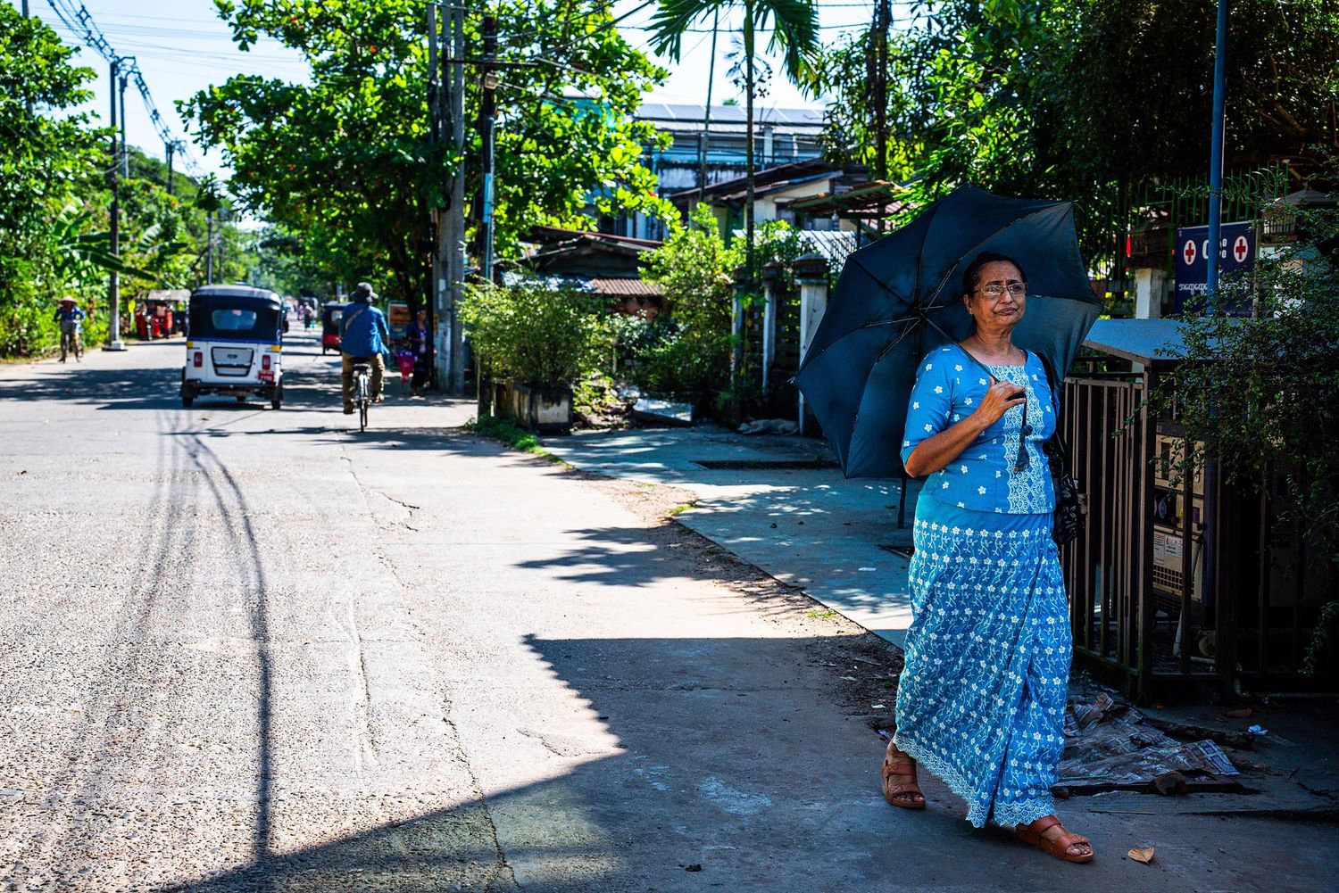 Person with umbrella walking down a sunny street in the Philippines with palm trees and houses in the background.